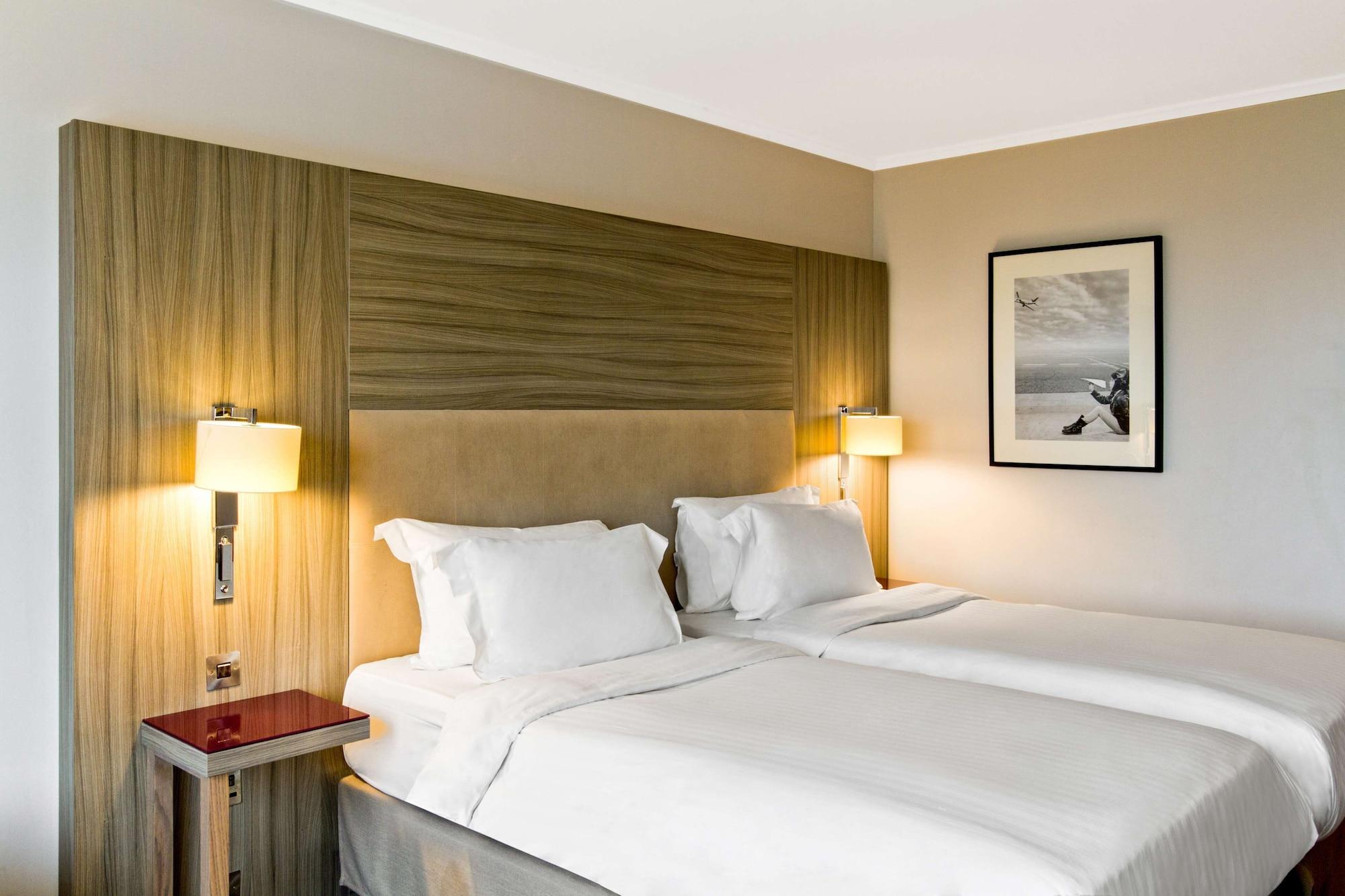 Radisson Blu Manchester Airport Hale  Esterno foto The photo shows a modern hotel room featuring two single beds with white bedding. The headboard is a combination of wood and fabric. There are bedside lamps on either side of the beds, providing soft lighting. On the wall, there is a framed black and
